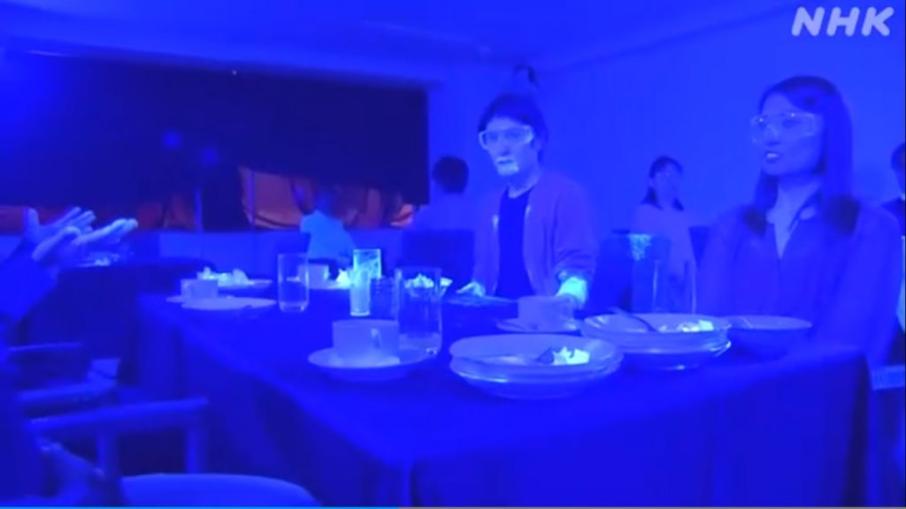 Imagem com luz ultravioleta: numa mesa de restaurante, pó fluorescente epslhado pela boca e pulso de um homem, por pratos e copos.