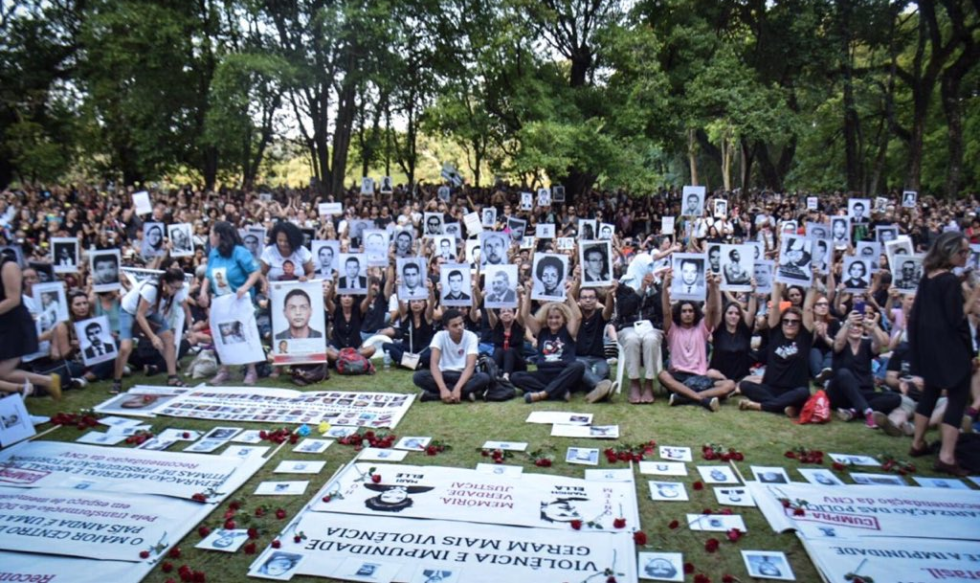 Centenas de pessoas sentadas em um gramado, muitas segurando cartazes com fotos de vítimas da ditadura. Sobre o gramado, algumas faixas e fotos deitadas.