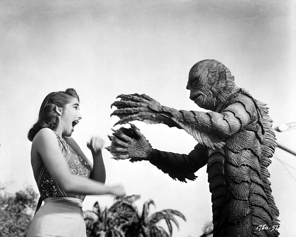 Foto de cena PB de 'O monstro da Lagoa Negra': o monstro, à direita, estendendo os braços e encarando uma mulher, à esquerda, que grita.